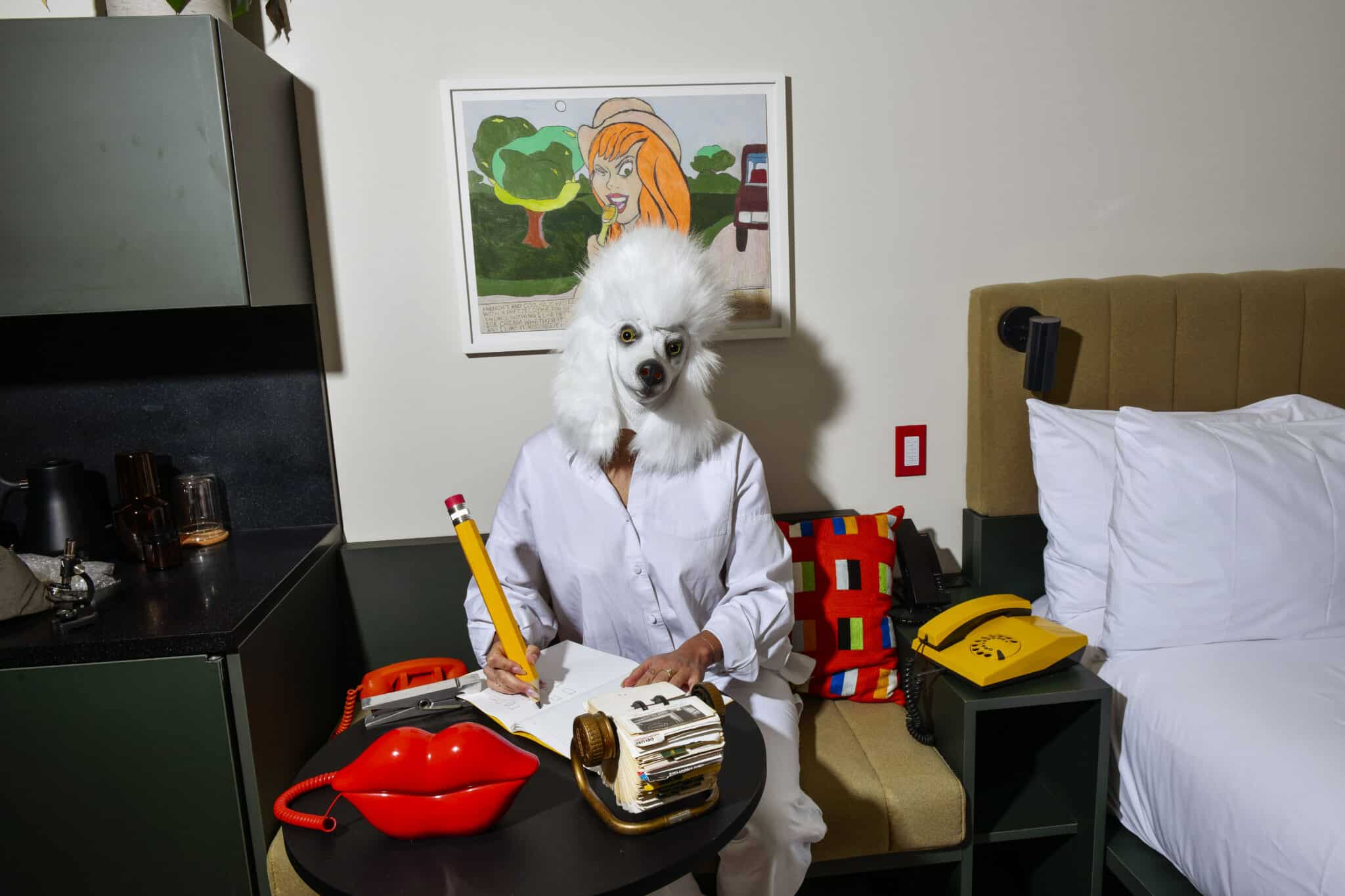 A person wearing a white poodle mask sits at a table in a hotel room, writing in a notebook.